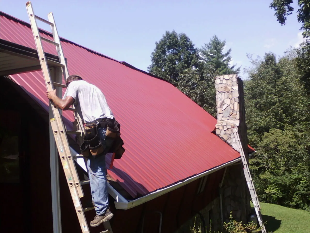 Roofer on a ladder beside a red metal roof and stone chimney — Soffit & Fascia Repair by Roofing Brothers in Ophiem